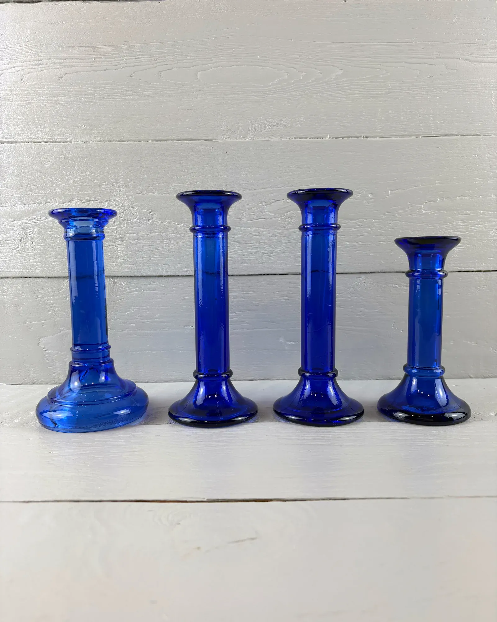 Set of four cobalt blue glass candlesticks in graduated heights, front view on white painted boards