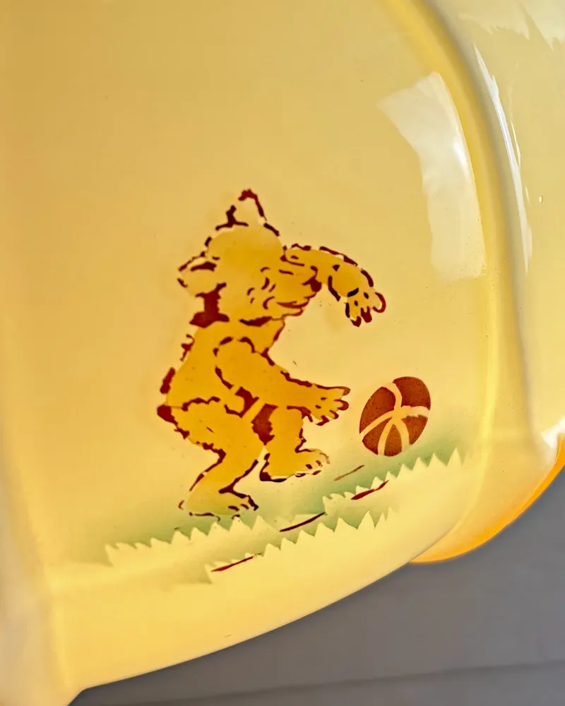 Detail of a printed bear kicking an orange ball on a strip of green grass, on glossy cream opal glass