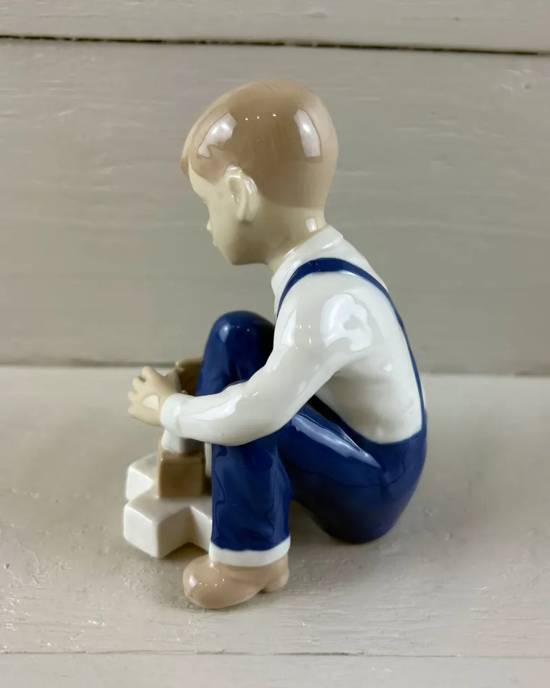 Side profile of a porcelain boy crouching over building blocks, blue dungarees and white shirt