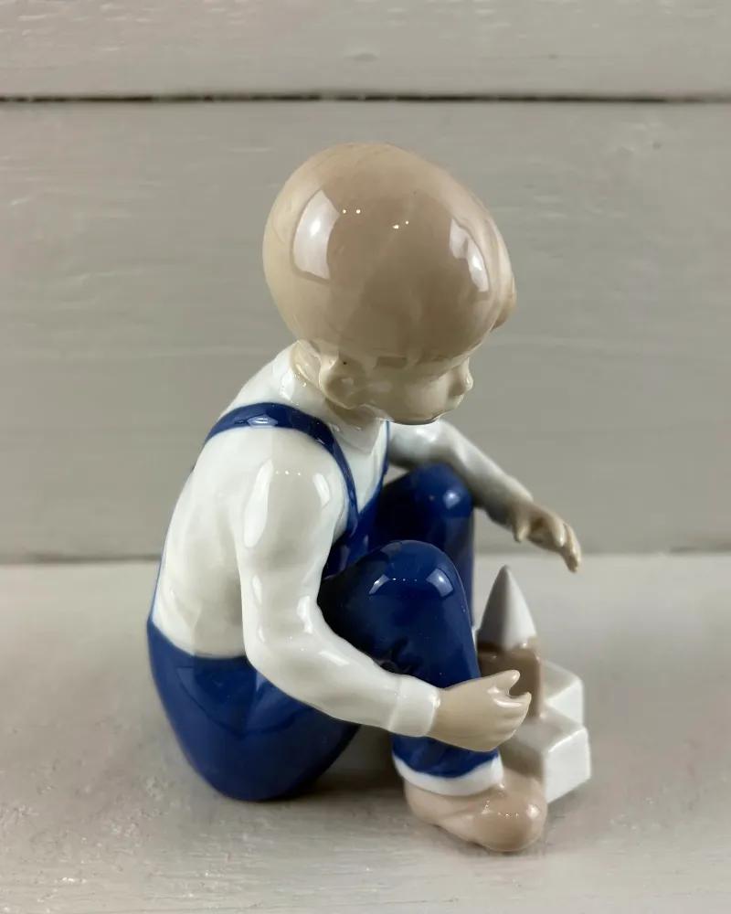 Porcelain boy figurine from the back right, reaching towards a pointed block, blue dungarees