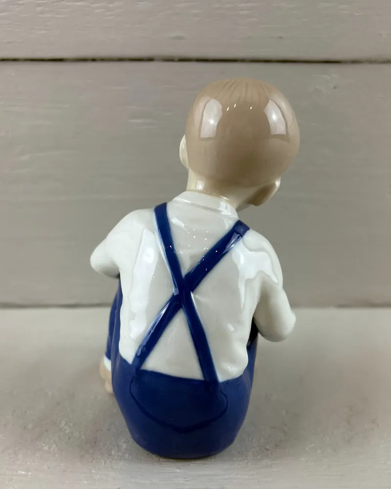 Back of a porcelain boy figurine showing the crossed blue straps of his dungarees over a white shirt