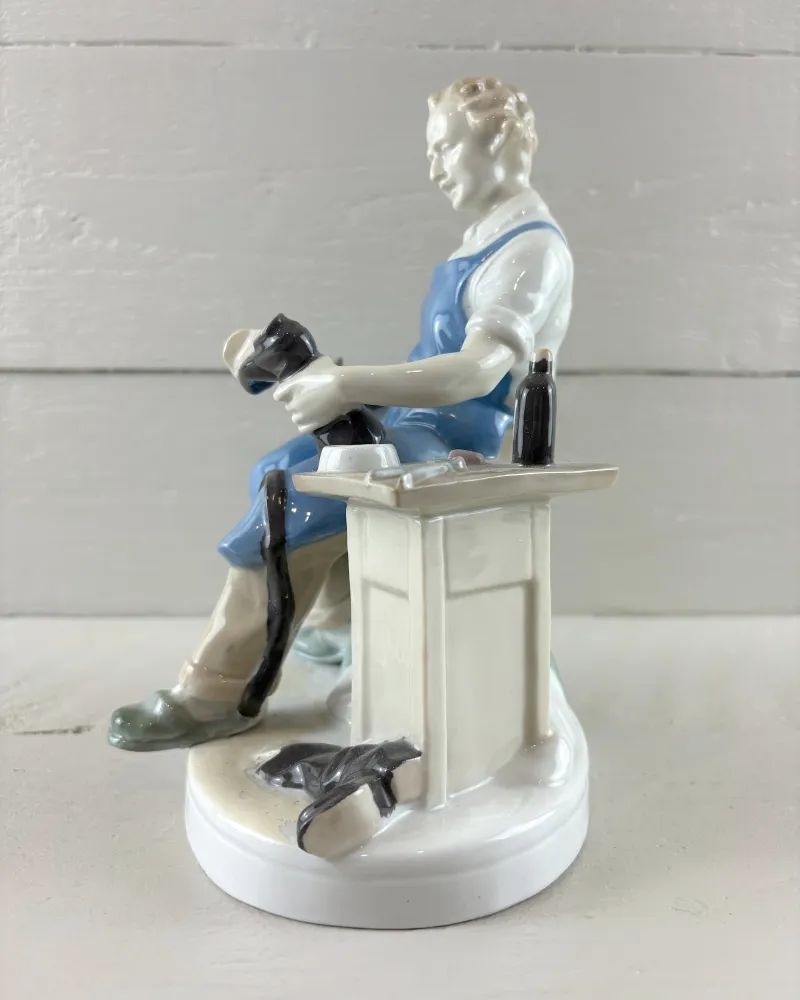 Right side of the porcelain cobbler figurine with workbench, dark bottle, bowl and black boots on the base