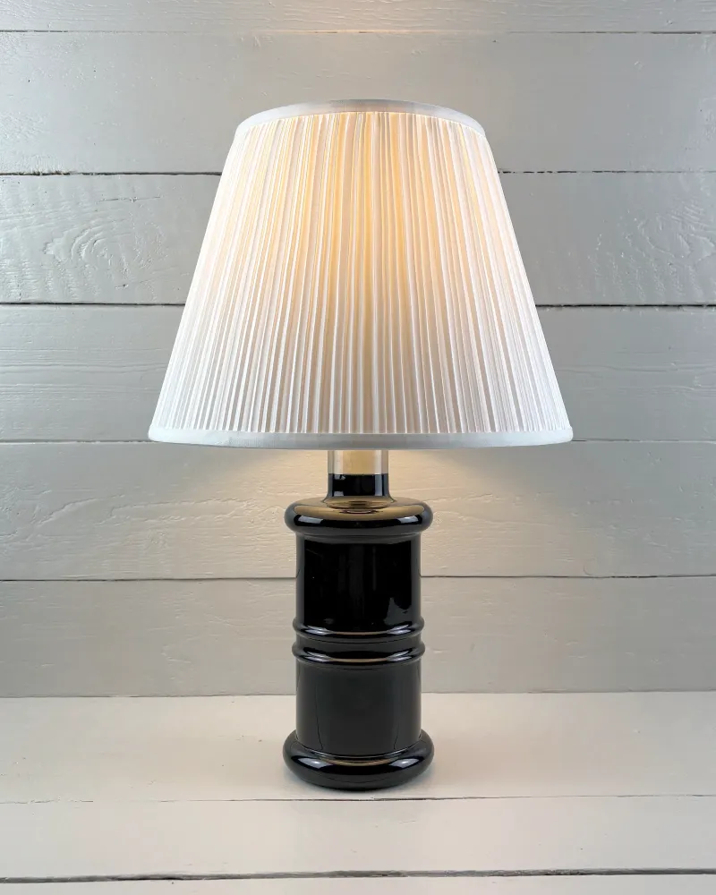 Holmegaard Apotheker black glass table lamp lit, with a pleated shade shown for illustration only