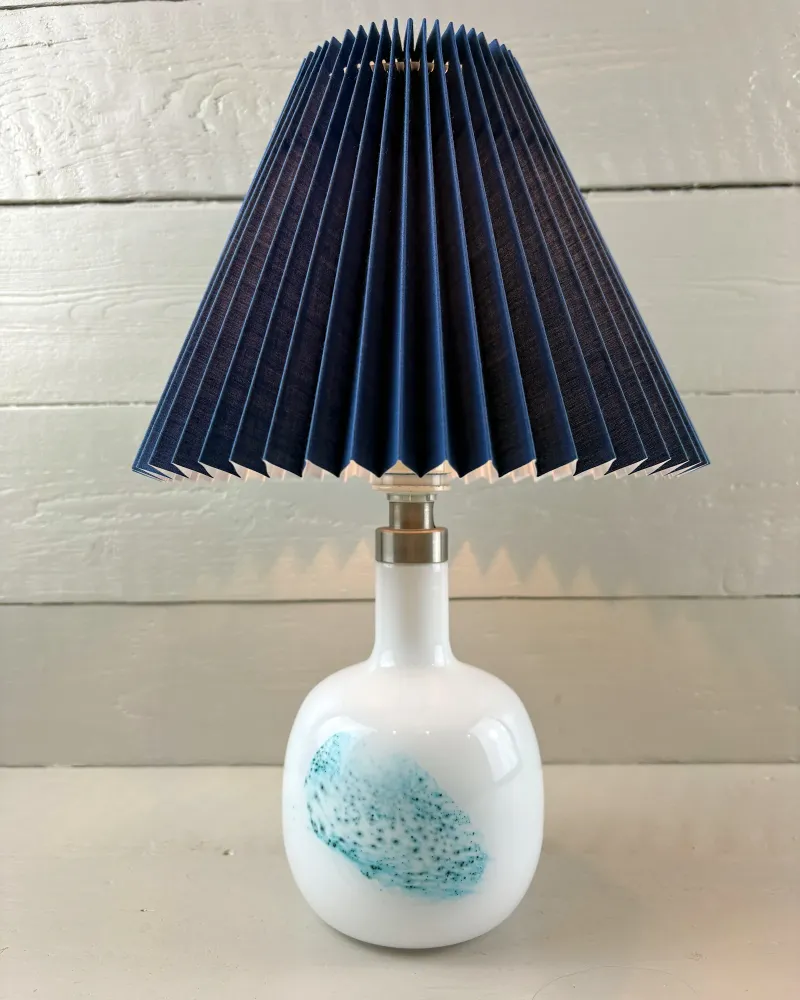 Holmegaard white opal glass table lamp switched on, soft light under an illustrative pleated shade