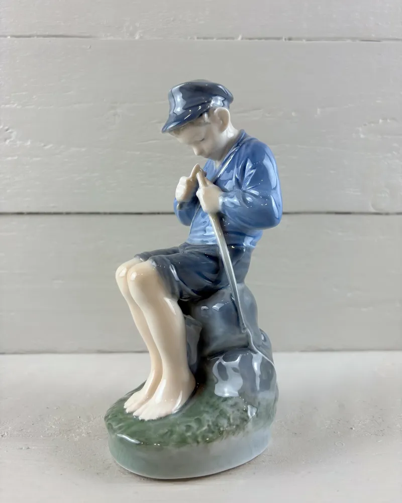 Side profile of Royal Copenhagen porcelain boy whittling a stick, showing the rock seat and grassy oval base