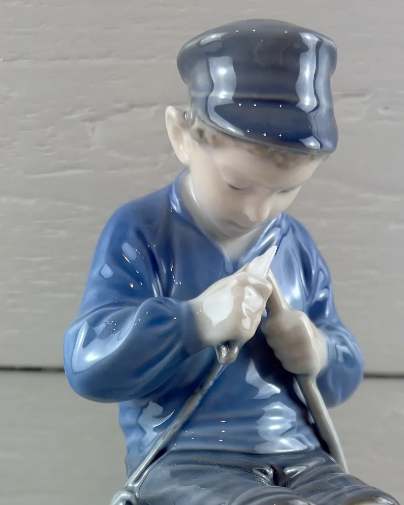 Close-up of the Royal Copenhagen Whittler's face, flat cap and hands carving a stick under glossy blue glaze