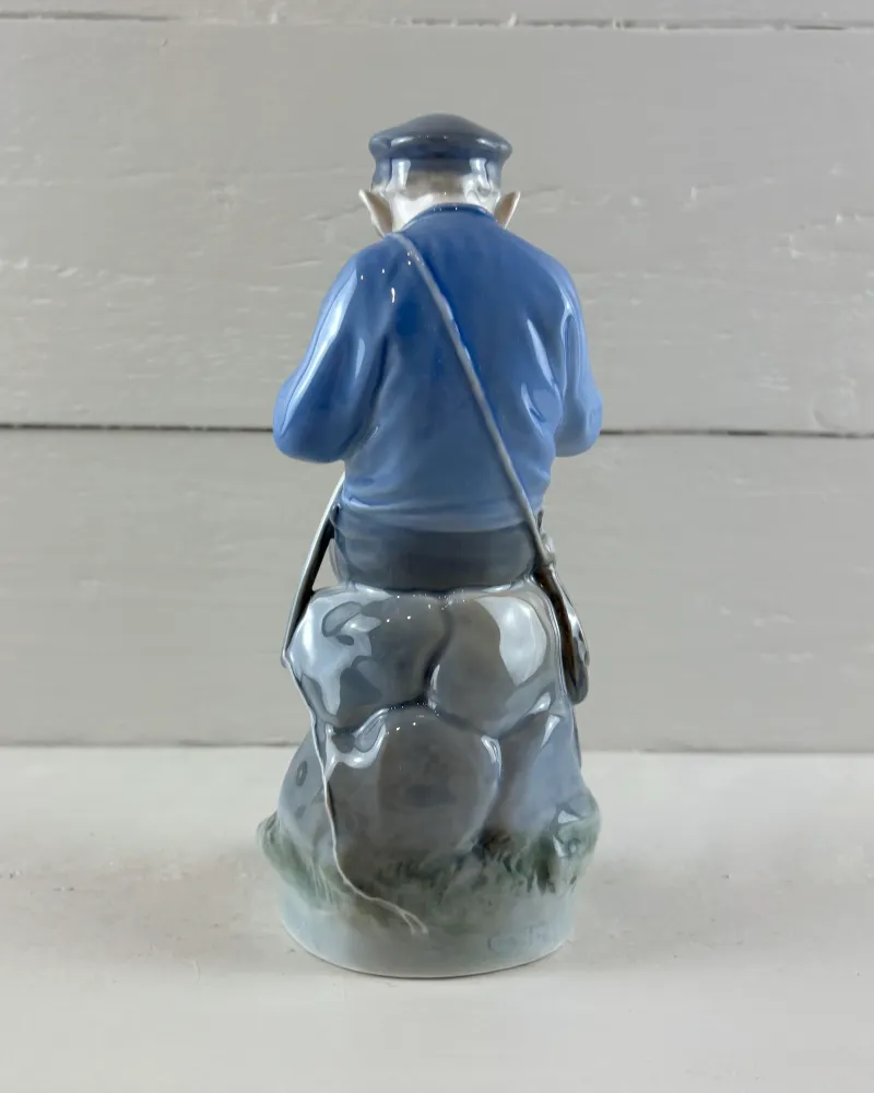 Back view of Royal Copenhagen porcelain boy figurine with a satchel strap across his blue jacket, seated on rocks
